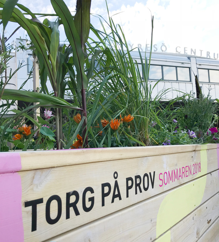 Texten "Torg på prov, sommaren 2018" tryckt på parkbänk. Grönska och Tyresö centrum-skylten i bakgrunden.   