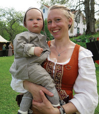 En glad mamma i medeltidskläder håller ett litet barn i famnen ute på Lilla Tyresö. Prinsvillan skymtar i bakgrunden.