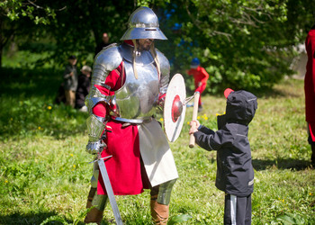 En medeltida riddare i rustning, svärd och sköld leker svärdskamp med ett litet barn.