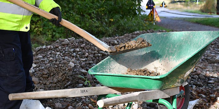 Person lastar med spade i grus i en skottkärra. 